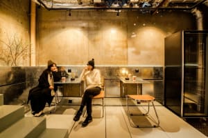 Two women sit in a dimly lit art gallery or indoor space with chairs, cups, and books, creating a cozy and intimate atmosphere
