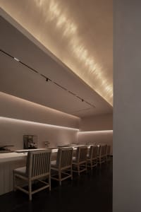 A dimly lit, modern restaurant with a long wooden dining table and metal/plastic chairs, creating an intimate ambiance