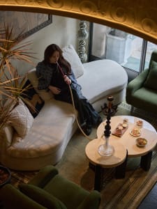 Cozy living area with a couch, chair, dining table, and a woman sitting on the couch smoking a pipe, surrounded by bowls and decorative items