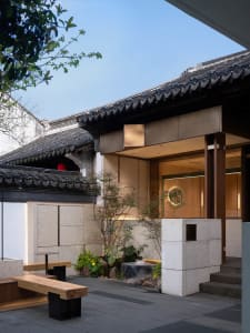 A traditional Asian-style house with a wooden exterior, stone wall, and red roof, featuring potted plants and a dining table in a cozy living space