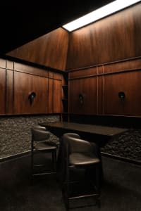 A modern restaurant or bar area with wooden walls and floors, featuring two black leather chairs and a marble dining table, well-lit with natural or artificial light