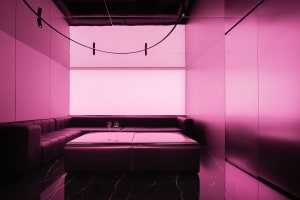 Modern restaurant with pink walls, a black couch, and a dining table, featuring a large window and wine glasses, creating a stylish atmosphere