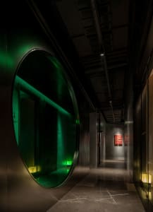 Industrial hallway with a circular mirror, concrete walls, and a painting on the wall, lit by two lights