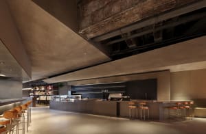 Open modern restaurant with concrete walls, gray accents, and overhead lighting, featuring a large counter and various seating options