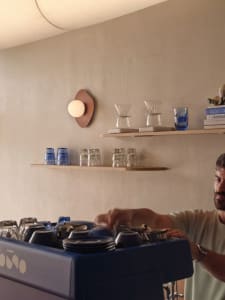 A man stands in a kitchen surrounded by cups, bowls, bottles, and books on various surfaces, creating a lived-in environment