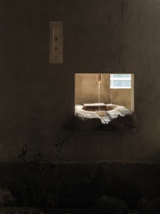 A serene stone courtyard with a fountain featuring a rock basin and two water spouts, surrounded by a rustic stone building