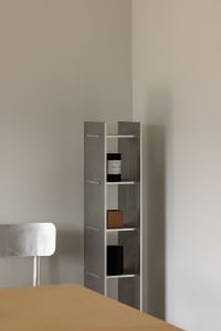 A modern room with a silver metal bookshelf featuring three shelves with books and bottles, showcasing a minimalist design with a focus on functionality and organization