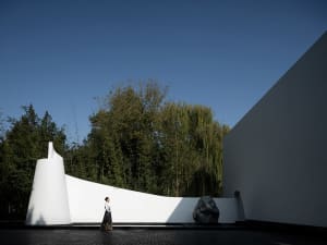 A woman stands in front of a modern building with a mirror, surrounded by a calm and serene environment with two large rocks nearby