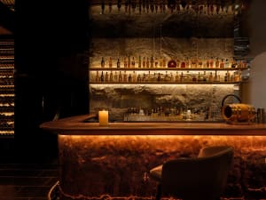 A sophisticated bar area with a long wooden counter, bottles of liquor, wine glasses, and two chairs, creating a comfortable environment for enjoying a drink