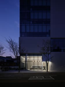 A modern office building at night with a tall white structure and many windows, featuring two people standing outside