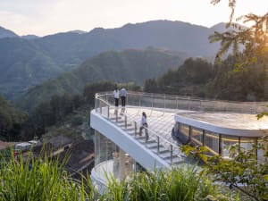 A modern building with a glass staircase and balcony on a mountain side, surrounded by greenery and mountains in the background