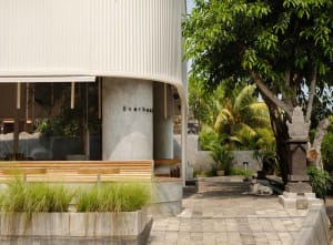 A modern building with a white exterior featuring a large circular structure, surrounded by potted plants and a dog sitting on the ground