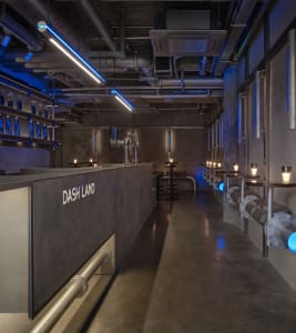 Dimly lit industrial bar area with concrete walls, a countertop, sinks, and numerous bottles
