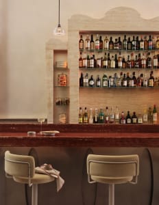 A cozy bar scene with a long wooden counter, two chairs, and bottles of liquor on display