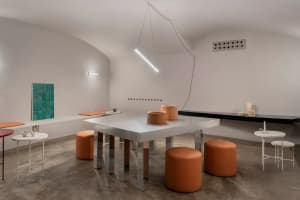 A modern cafe with a white table and orange stools, surrounded by chairs and natural light from windows