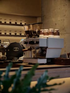 A cozy coffee shop interior with a white espresso machine, cups, and bottles on a countertop, creating a welcoming atmosphere for customers