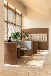 A restaurant interior with wooden walls and floors, natural light, and potted plants, featuring a person sitting at a bench