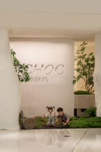 Two children sitting on rocks in front of a white wall with a sign, surrounded by potted plants and a chair in a casual outdoor setting