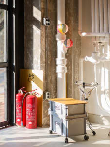 A cluttered living area with a window, red fire extinguishers, bottles, and a bowl, suggesting a functional yet somewhat disorganized space