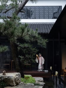 A serene courtyard setting with a woman in traditional attire standing in front of a building, surrounded by trees and several chairs, conveying a sense of cultural significance and tranquility