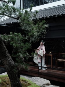 A woman in traditional attire stands in front of a building with a wooden table and chairs, surrounded by potted plants, conveying a sense of tradition and cultural significance