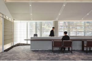 A modern office reception area with a gray carpet, white walls, and a reception desk with two people seated at it, surrounded by two chairs and a clean, professional atmosphere