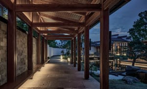 A calm and inviting outdoor space with wooden pillars supporting a roof, featuring benches along the walkway