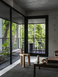 Modern living space with concrete walls and flooring, featuring a large window overlooking a forest, two chairs, a dining table, and a potted plant