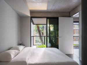 A modern bedroom with a white bed in front of a window, surrounded by concrete walls, potted plants, and a chair, creating a calm and inviting atmosphere