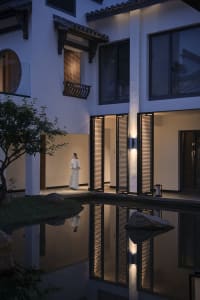 A woman walks across a bridge in front of a large Asian-style building with white walls and multiple windows and doors, surrounded by potted plants