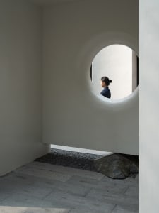 A woman stands in front of a concrete wall with a round window, surrounded by a calm and contemplative atmosphere