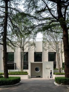 A modern building with a large circular window, surrounded by trees and featuring white walls and a gray roof