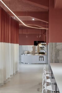 A modern restaurant dining area with a large wooden table surrounded by chairs, cups, and a kitchen with a refrigerator and oven in the background