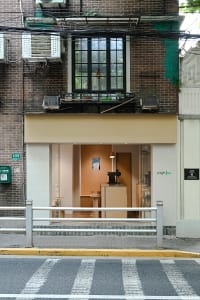 A small storefront with a wooden door and window, two chairs, and brick walls, creating a warm and inviting atmosphere with natural light