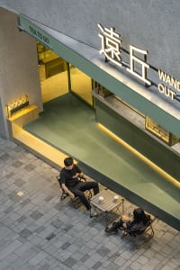 A modern Asian restaurant with green walls and a glass door, featuring a relaxed and inviting atmosphere with two people enjoying drinks outside