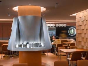 A casual restaurant dining area with modern decor, featuring tables, chairs, and a large silver bowl on display, with people sitting and standing around the tables