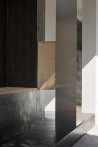 Black building with a wooden wall and a glass wall, featuring three pillars and a window allowing natural light to enter the space