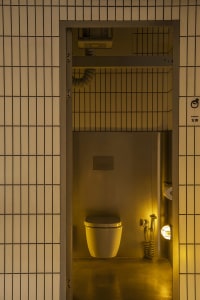 A functional bathroom with two metal sinks, a white toilet, and a mirror above one of the sinks, set against a yellowish background
