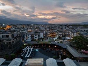 A cityscape at dusk featuring a large building with a mix of residential and commercial spaces, stone and glass facade, and a vibrant urban atmosphere