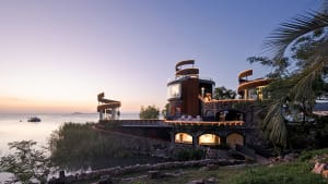 A large house on a hillside overlooking water, with a stone exterior, multiple levels, and a circular structure, featuring outdoor seating areas and a luxurious atmosphere