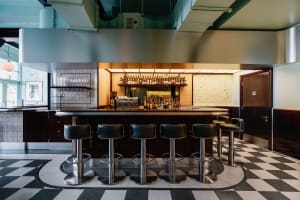 A casual bar area with a long wooden counter and multiple stools, surrounded by chairs and wine glasses, with a variety of bottles on display