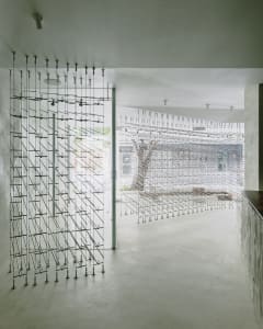A modern open-plan living area with a kitchen, featuring a wire mesh door, a tree trunk, and a minimalist atmosphere