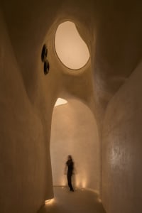 A woman walks down a well-lit, curved concrete hallway with two clocks on the wall, one near the left side and one further back