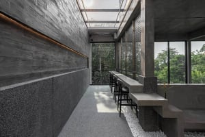 A modern restaurant with a large dining table, concrete walls, a glass ceiling, and potted plants, creating an inviting atmosphere for socializing and dining