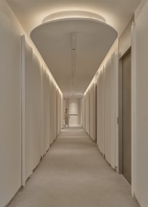 A long, modern hallway with white walls and floors, featuring several doors and a person standing in an adjacent room