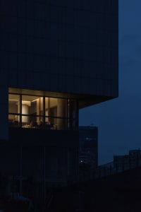 A modern high-rise building at night with a glass structure, multiple windows, and well-lit interior, featuring chairs and a sophisticated atmosphere