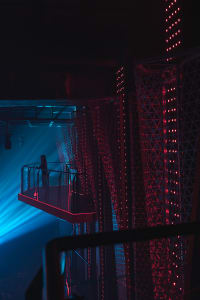 A woman stands on a balcony looking down at the area below, surrounded by a residential or commercial space with a sense of curiosity and contemplation
