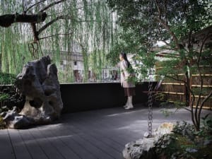 A serene garden area with a woman in a white dress standing near a large rock, surrounded by potted plants and two chairs, with a peaceful atmosphere