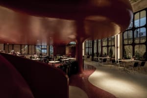 A modern restaurant dining area with multiple tables, chairs, and potted plants, surrounded by red walls and natural light
