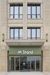 A modern building with a green sign reading M Stand, featuring multiple windows, two clocks, and a contemporary urban atmosphere